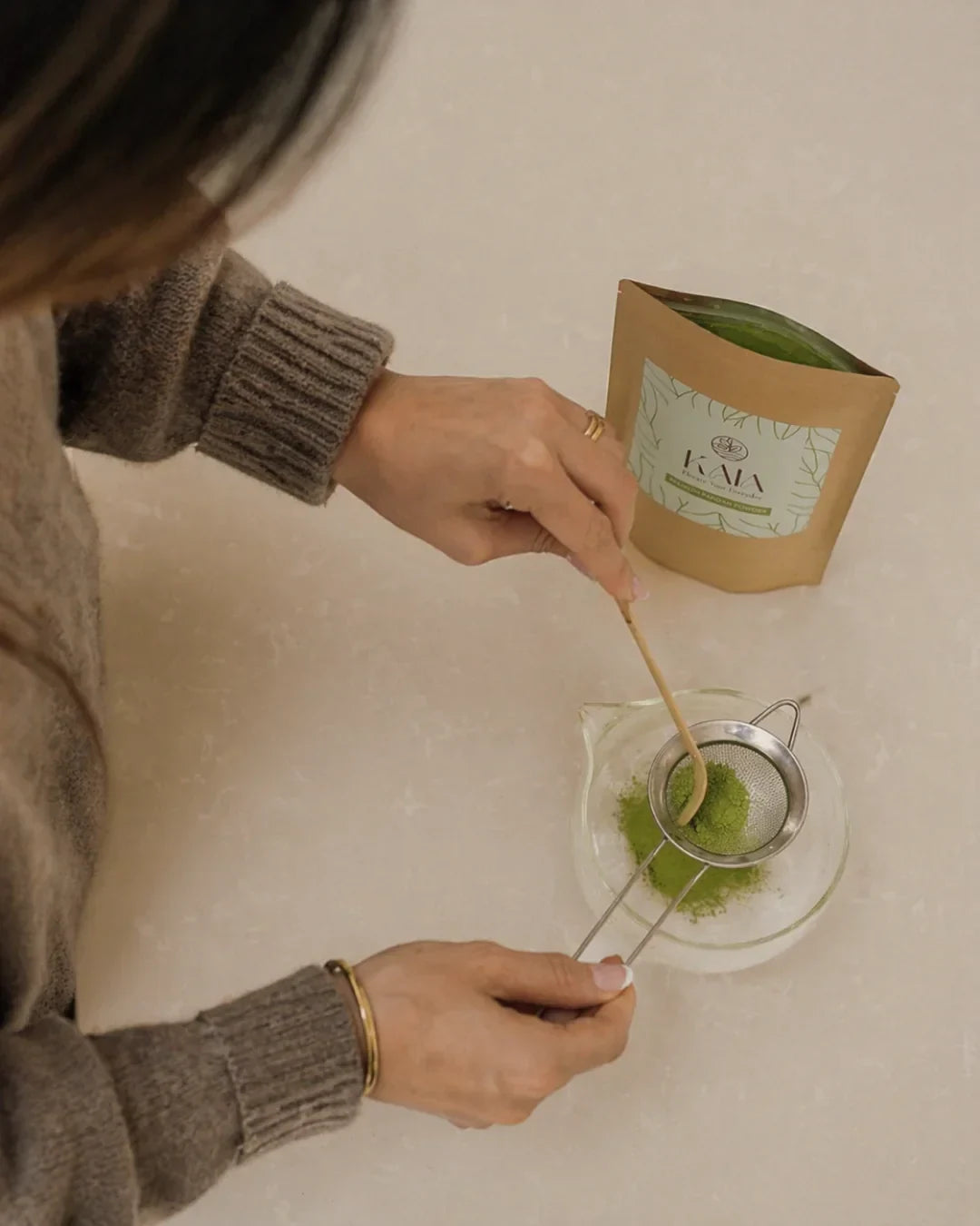 Top-down view of hands sifting KAIA Origin matcha powder through a fine sieve into a glass bowl, kraft pouch in background